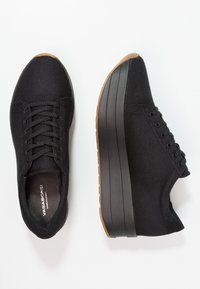 Pari mustia korokepohjasneaker-pareja, joissa on teksturoidusta kankaasta yläosat, mustat nauhat, paksut mustat välipohjat sekä ruskeat kumiset varvas- ja pohjavarustukset.