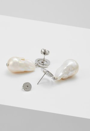 Par de pendientes de plata con perno, cada uno con una gran perla barroca blanca irregular sobre una superficie blanca lisa.