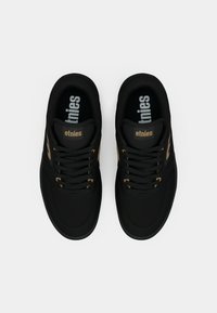 Zapatillas negras con una parte superior de tela suave, que cuentan con detalles dorados y cordones negros. Las plantillas muestran el nombre de la marca "etnies" en blanco.