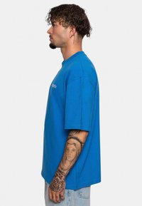 Blaues oversize T-Shirt aus Baumwolle, mit rundem Ausschnitt, kurzen Ärmeln und einem schlichten Logo auf der Brust. Das Modell hat Tattoos am Arm.