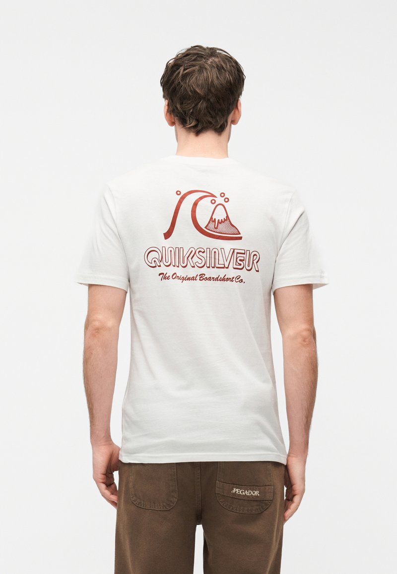 Άνδρας με καστανά μαλλιά που φοράει λευκό μπλουζάκι Quiksilver με κόκκινο λογότυπο κύματος και βουνού, και καφέ παντελόνι Pegador, στέκεται με την πλάτη γυρισμένη.