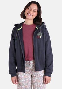 Haut à zip marin avec des accents verts et un design brodé sur la poitrine, associé à un t-shirt bordeaux et un pantalon à motifs floraux. Texture de tissu douce.
