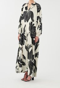 Robe maxi en tissu léger avec un fond crème et de grands motifs floraux noirs. Manches longues avec poignets élastiques, détail noué au col.