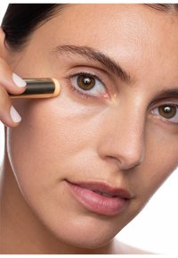 Correttore in stick con un rivestimento in metallo dorato, applicato sotto l'occhio su una pelle chiara e levigata, evidenziando un look trucco naturale.