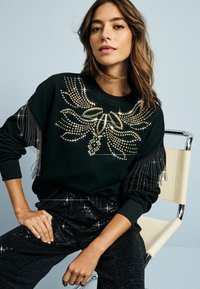 Felpa nera con design arricchito da borchie dorate e argentate, con dettagli di frange sulle maniche. Abbinata a pantaloni neri brillanti.