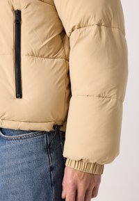 Beige puffer-takki, jossa on vetoketjullinen tasku, paksu vuoraus, joustavat hihansuut ja kiristettävä helmavarsi, yhdistettynä sinisiin farkkuihin.