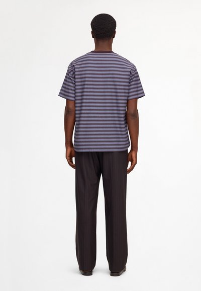 Homme debout de dos, portant une chemise rayée violet et noir à manches courtes et un pantalon ample sombre à fines rayures.