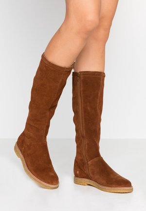 Brune ruskinds knæhøje støvler med sidelås, båret på bare ben, stående på en hvid reflekterende overflade.