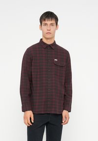 Camicia a quadri rossa e nera con colletto a bottoni, maniche lunghe, taschino singolo sul petto e etichetta del marchio sulla tasca. Tessuto strutturato.