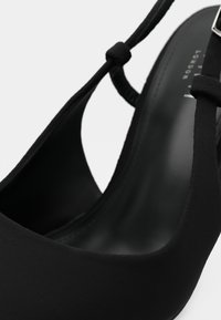 Μαύρο πέδιλο με τακούνι τύπου slingback, με λείο, ματ φινίρισμα, που διαθέτει στρογγυλεμένη μύτη και λεπτό λουράκι στερεωμένο με μικρή ασημένια αγκράφα.