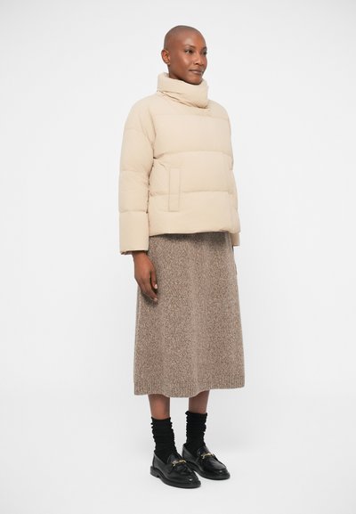 Beige puffer jacket with a high collar, short sleeves, and a front pocket; paired with a knee-length textured brown skirt and black shoes.