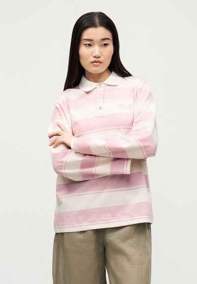 Νεαρή γυναίκα με μακριά σκούρα μαλλιά, που φοράει ένα ροζ και λευκό ριγέ polo με μακριά μανίκια και μπεζ παντελόνι, με σταυρωμένα χέρια, κοιτάζει πλάγια.