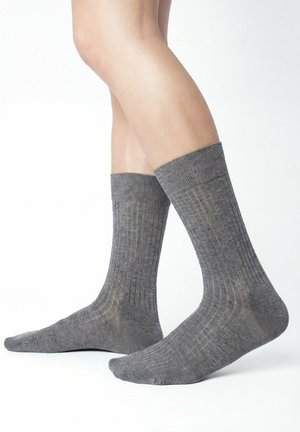 Gambe con calzini grigi a coste di media lunghezza fino al polpaccio, in piedi su una superficie bianca con un piede piatto e uno leggermente sollevato.