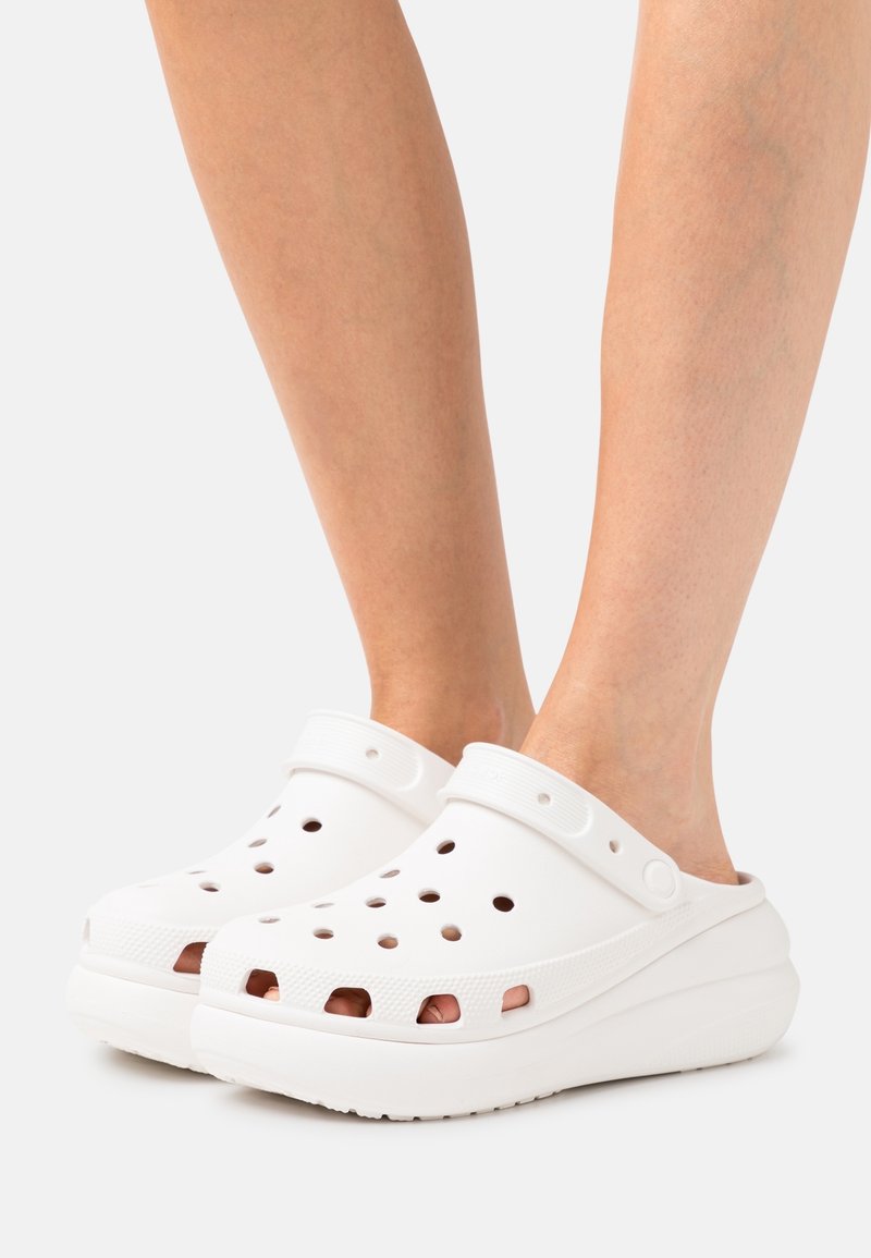 Personne portant des sabots blancs à enfiler avec des trous d'aération et des brides réglables au talon sur un fond clair.