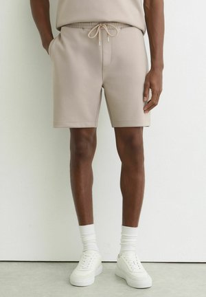 Dorosły ubrany w beżowe szorty z troczkiem, białe skarpetki sportowe i białe trampki, stoi na tle gładkiej białej ściany.