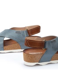 Sandalias de cuero azul con correas cruzadas, correa ajustable de color marrón en el talón, suela de cuña de corcho y suela blanca. Logotipo de la marca en el lateral.