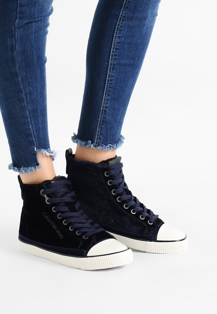 Jalkoja, jotka pukeutuvat tummansinisiin samettisiin korkea-ankkalippusiin, joissa on valkoiset kumiset varvaskärjet ja pohjat, yhdistettynä risaisiin skinny-farkkuihin.