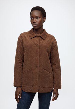 Femme aux cheveux courts portant une veste matelassée marron avec des poches avant et un jean bleu foncé sur un fond clair uni.
