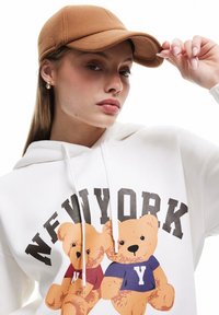 Sudadera blanca con osos de dibujos animados y texto "NUEVA YORK", combinada con una gorra marrón. Tejido suave con una textura blanda.