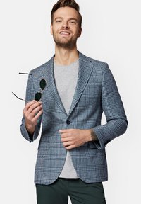 Niebieska marynarka w kratę z wyraźnymi klapami, zestawiona z szarym T-shirtem. Mężczyzna trzyma czarne okulary przeciwsłoneczne, prezentując nowoczesny, dopasowany krój.