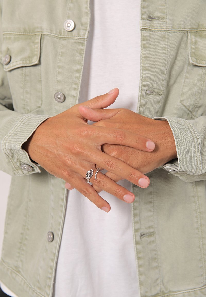 Anelli d'argento esposti sulle mani, caratterizzati da un design a forma di tartaruga e accenti texture, abbinati a una giacca di jeans verde chiaro sopra una t-shirt bianca.