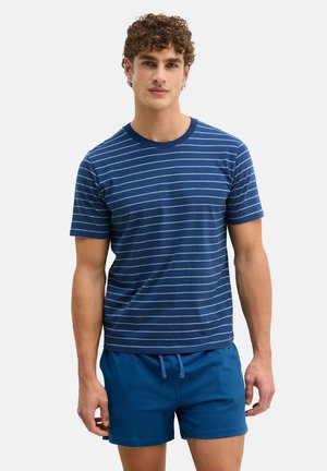 Junger Mann mit lockigem Haar, der ein marineblau gestreiftes T-Shirt und einfarbige blaue Shorts vor einem einfarbigen hellen Hintergrund trägt.