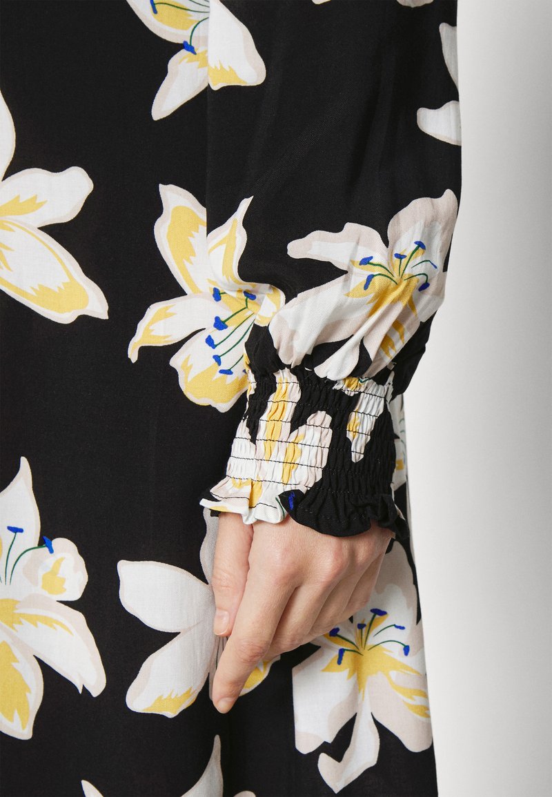 Vestido de tecido preto com grandes padrões florais brancos e amarelos. A manga tem detalhes franzidos com bordas em babados.