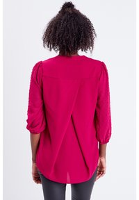 Blusa fucsia con mangas abullonadas texturizadas y un diseño trasero fluido. Fabricada en tela ligera, presenta sutiles pliegues y un dobladillo redondeado.