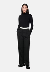 Woman in black turtleneck and wide-leg black pants with white waistband, hands in pockets, standing against a plain white background.