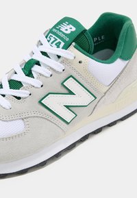 Baskets New Balance 574 en daim beige et mesh blanc avec des accents verts, lacets blancs et grand logo "N" sur le côté.
