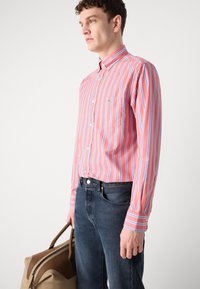 Camisa de rayas en rojo, azul y blanco, con cuello abotonado y un bolsillo en el pecho, combinada con jeans azul oscuro y un bolso marrón.