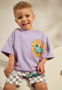 Lila übergroßes T-Shirt mit "Hot Wheels"-Grafik, kombiniert mit grau-weiß karierten Shorts mit türkisfarbenen Akzenten. Glatte Stoffstruktur.