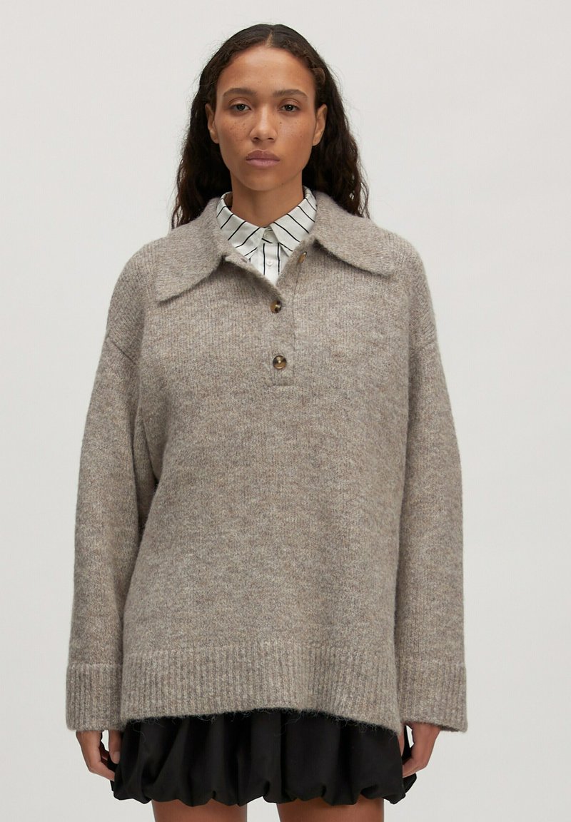 Beiger Pullover aus Wollmischung mit Kragen, einer Drei-Knöpfe-Leiste und gerippten Bündchen. Getragen über einem gestreiften Button-Down-Hemd, was einen geschichteten Look erzeugt.