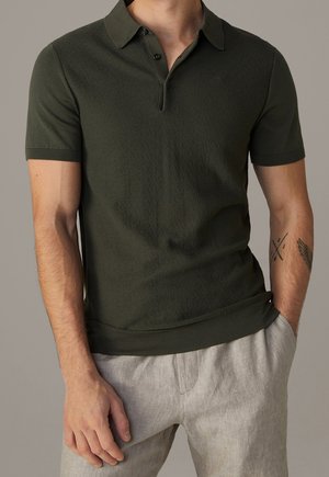 Mørkegrøn kortærmet polo skjorte lavet af tekstureret stof. Har en knaplukning og ribbede manchetter. Parret med lysegrå bukser.