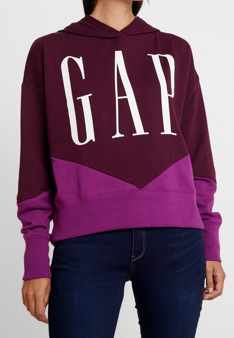 Felpa borgogna e viola con grande scritta bianca "GAP", caratterizzata da un design a blocchi di colore e un cappuccio con coulisse. Texture morbida e vestibilità rilassata.