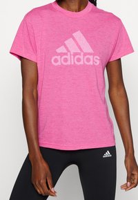Camiseta deportiva rosa jaspeada de manga corta, con un gran logo blanco de adidas en el frente. Combinada con leggings negros.