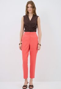 Brown sleeveless top with front buttons, paired with coral tapered trousers and brown sandals with bow detail. Smooth textures throughout.