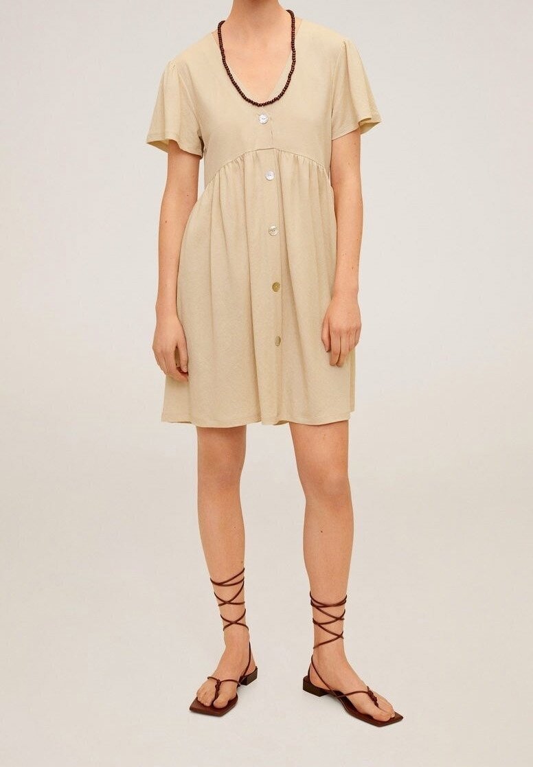 Femme portant une robe beige à manches courtes boutonnée sur le devant avec un col en V et des sandales plates marron à lacets.