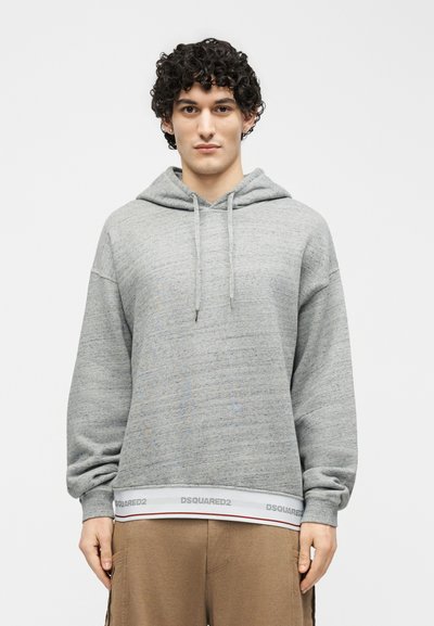 Giovane uomo con capelli ricci che indossa una felpa con cappuccio grigio chiaro con il dettaglio "DSQUARED2" sull'orlo e pantaloni marroni, in piedi davanti a uno sfondo bianco.