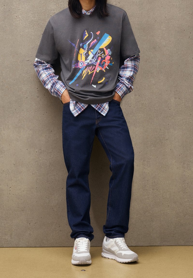 Grå grafisk t-shirt lagd över en blå rutiga långärmad skjorta, kombinerad med mörk denim jeans och grå-vita sportskor.