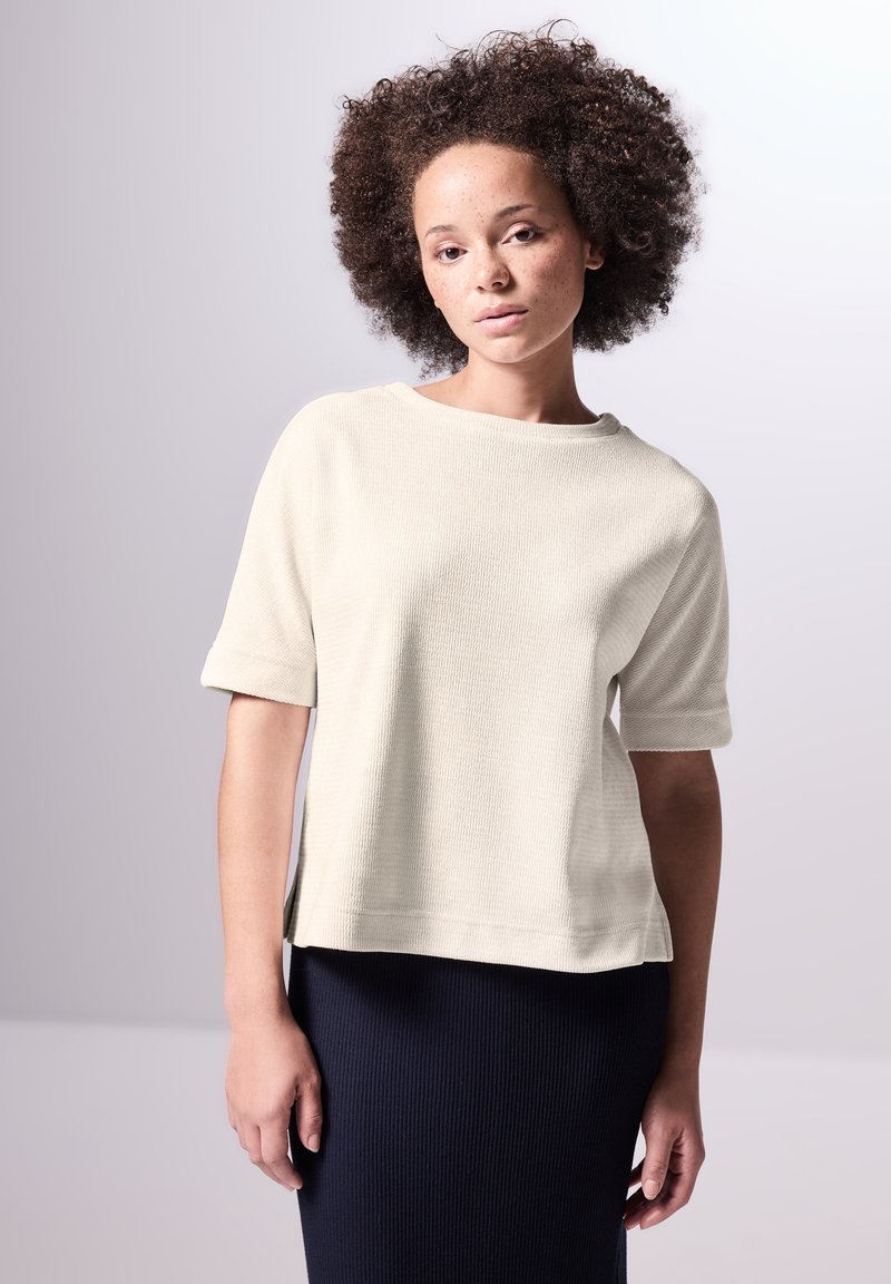 Licht beige textuur top met korte mouwen en een ontspannen pasvorm en ronde halslijn, gecombineerd met een donker geribbeld rokje, tegen een neutrale achtergrond.
