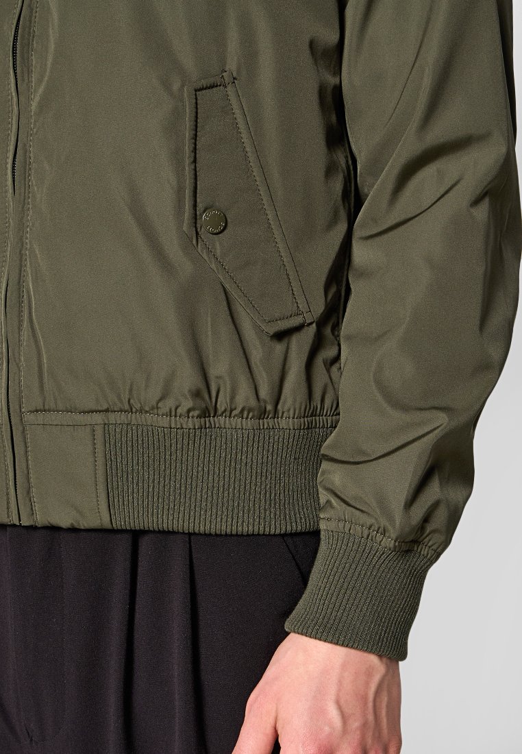 Veste bomber vert olive avec un ourlet et des poignets côtelés, fermeture éclair et poche arrivante avec détail à bouton-pression. Tissu lisse et léger.