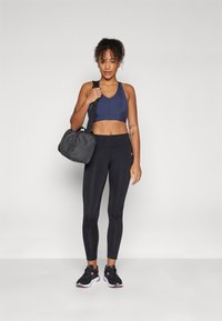 Mornarski športni nedrček, črne legice in črni tenisky s roza poudarki; nosi sivo športno torbo. Gladka tekstura in prilegajoča zasnova.