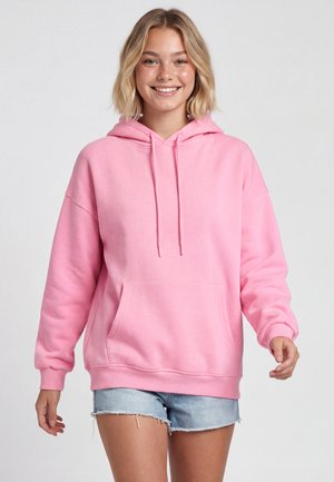 Ung kvinde med kort blondt hår, der smiler, iført en lyserød hættetrøje og lyseblå denimshorts mod en ensfarvet lys baggrund.
