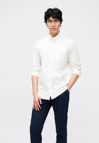 Bela srajca s poklopcem, dolgimi rokavi, zavihanimi do komolcev, v kombinaciji s temno modrimi hlačami. Gladek tekstura, klasičen kroj.