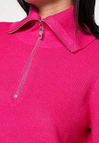 Top rosa fitted in maglia con una texture a coste e colletto con zip. Presenta una zip argentata lucida e uno scollo pulito.