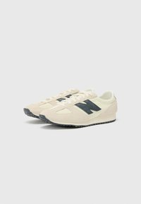 Beige suede sneakers with a black "N" logo, textured upper, cream accents, and rubber soles, featuring a classic lace-up design.