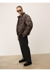 Vyras vilkintis per didelį rudą pūkinį švarką, juodas kelnes, juodus žygių batus ir saulės akinius, stovi prieš paprastą smėlio spalvos foną.