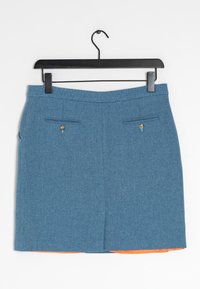 Falda azul con patrón en espiga y dos bolsillos abotonados en la parte trasera colgando de una percha de madera negra, con un forro naranja visible en el dobladillo.