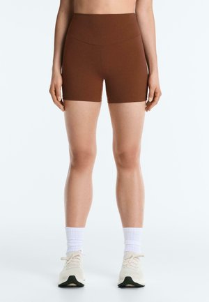 Personne portant un short de vélo marron taille haute, des chaussettes blanches classiques, et des chaussures de sport blanches, debout devant un fond clair uni.